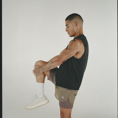 A man performing a knee hug stretch in a black tee and brown shorts.