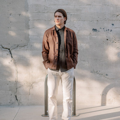 A man in a brown jacket and white pants stands in front of a concrete wall.