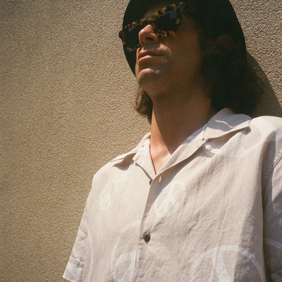 A man stands against a concrete wall wearing sunglasses, a black bucket hat and a shirt with white peace signs printed all over it.