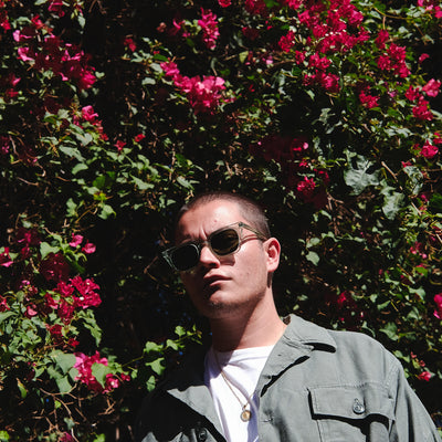 A man wearing sunglasses and an olive overshirt stands in front of pink flowers.