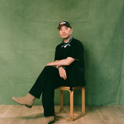 A man in a baseball hat and all black outfit sits on a wooden stool.