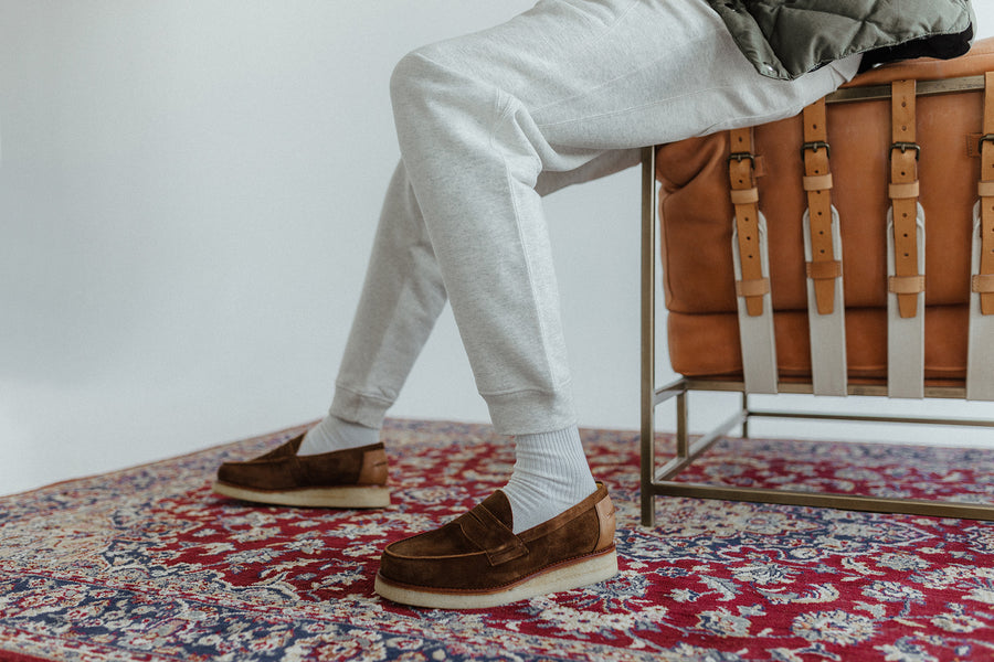 A pair of legs in sweatpants and brown loafers on a rug.