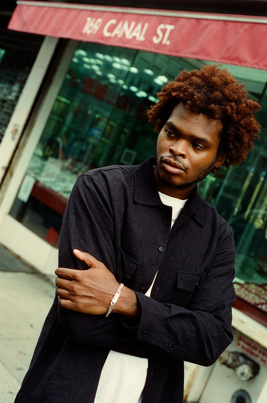 A man in a black shirt and silver chain necklaces stands outside.