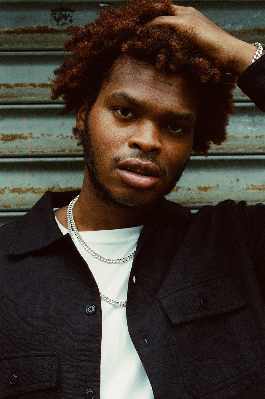 A portrait of a man in white gold chains stands in front of a metal gate.