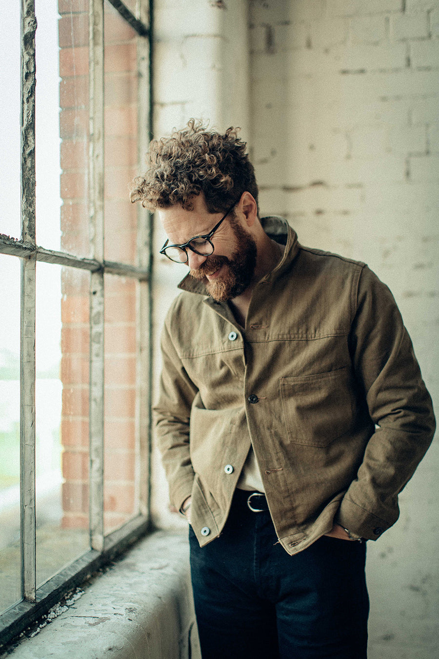 A man smiles at the floor and wears a tan jacket by a window.