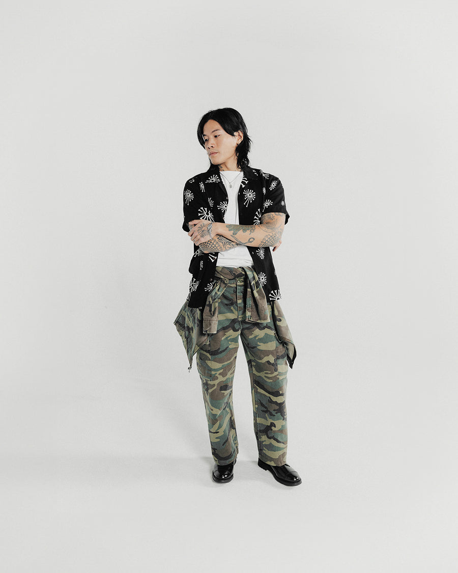 A man poses in a black short sleeve shirt and camo pants.