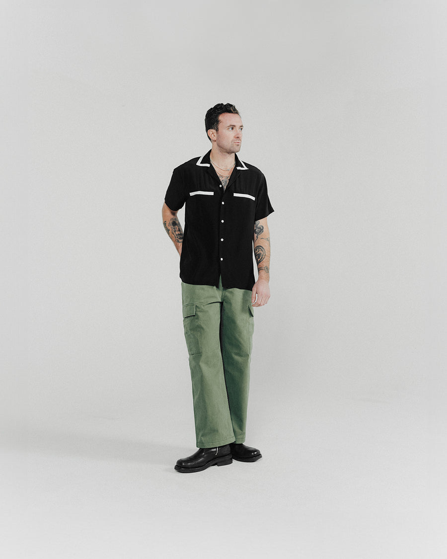 A man poses in a black shirt with white accents and olive wide leg pants.