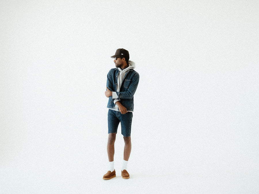 A man in a denim jacket and jorts stands on a white backgroud.