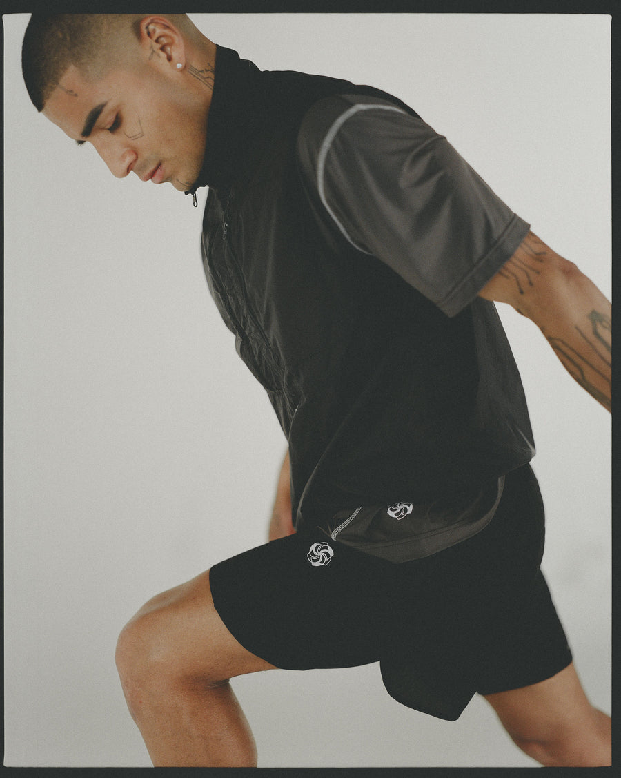 A man in a grey workout tee and black vest leans forward in a stretch position.