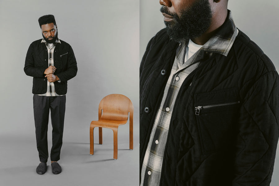 A man in a black liner jacket with a plaid shirt underneath stands in front of a wooden chair.