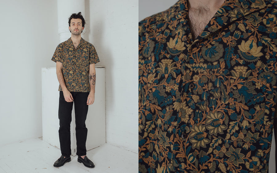 A man in a printed black shirt and black jeans looks into the camera.