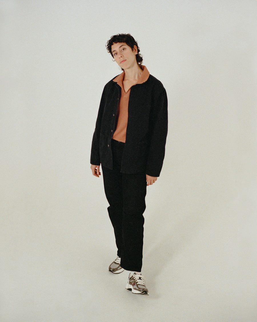 A woman in a reddish-orange knit polo under a black chore jacket poses.