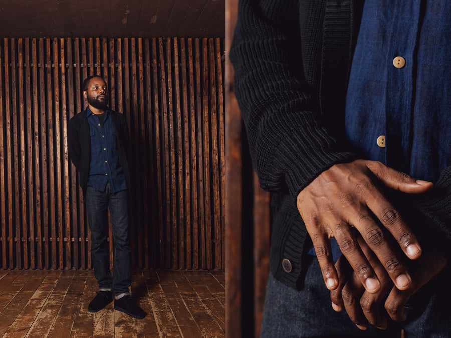 A man in a black sweater and indigo blue shirt stands in front of a wooden slat wall.
