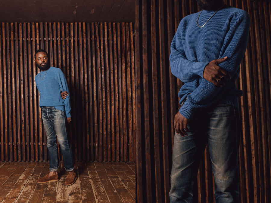 A man in a light indigo knit sweater stands in front of a wooden slat wall.