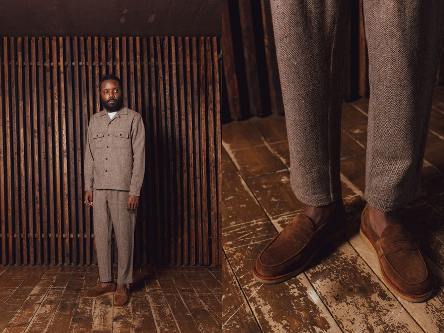 A man in a brown wool shirt and pants stands before a wooden wall.