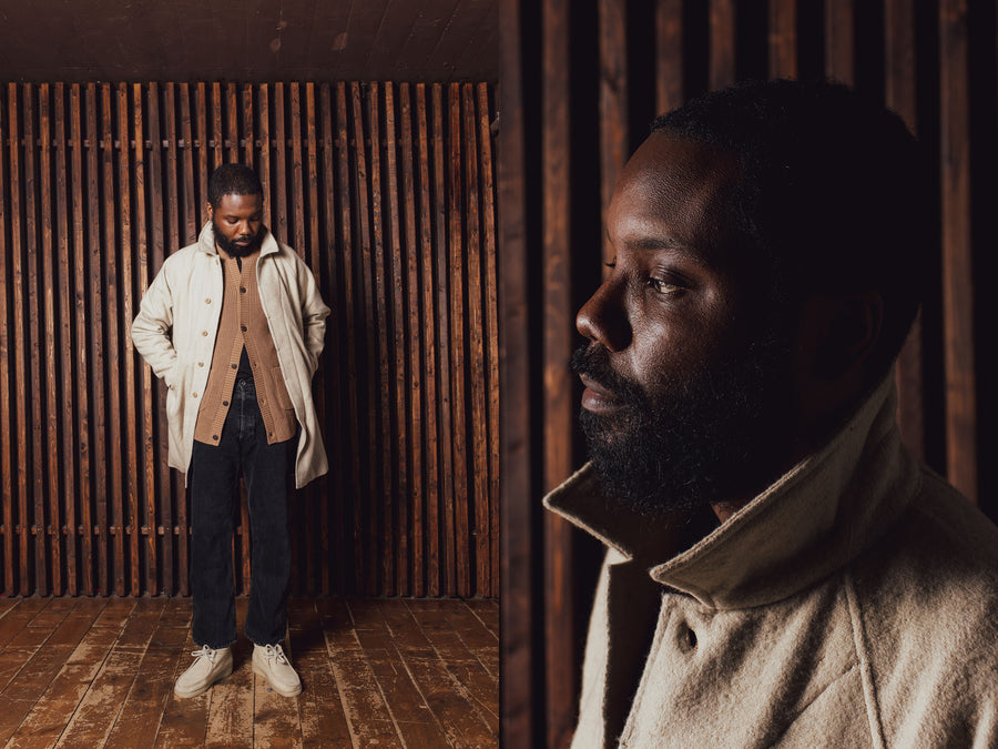 A man in a light tan overcoat with brown sweater and black jeans stands in front of a wooden slat wall.
