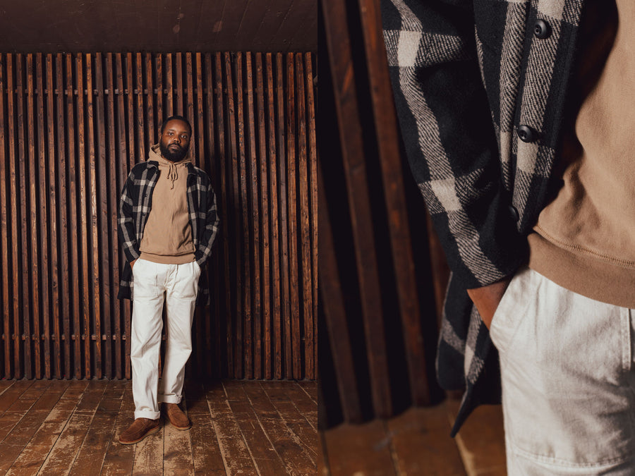 A man in a black plaid wool overcoat and tan hoody stands in front of a wood slat wall.