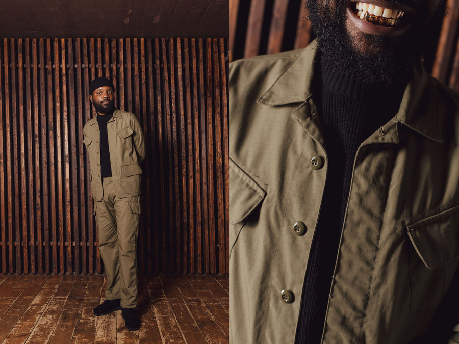 A man in a black sweater and full olive ripstop suit stands in front of a wooden slat wall.