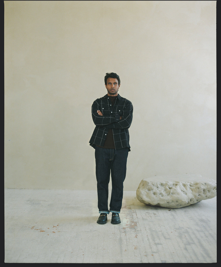 A man in a black windowpane patterned shirt and black pants stands next to a rock.