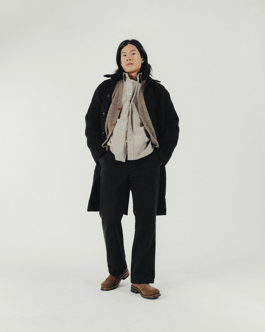 A man in a striped shirt and long wool coat and brown boots stands with hands in pockets.