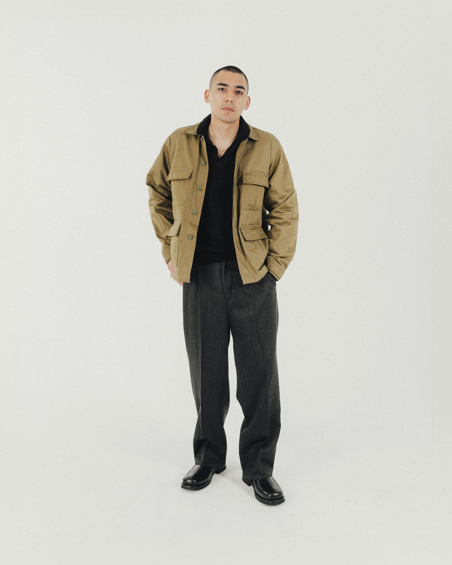 A man in a tan jacket and grey pleated pants stands with his hand in his back pocket.