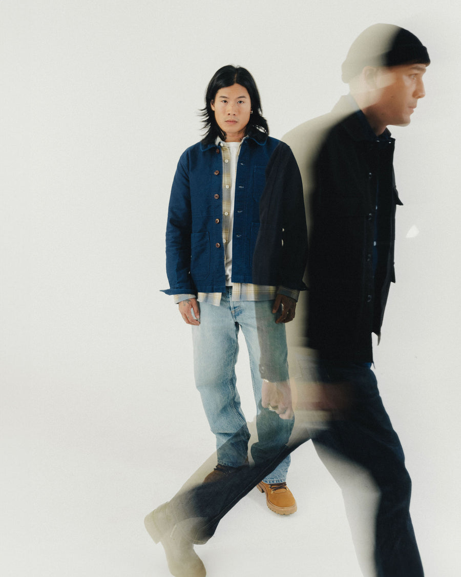 A man in a blue jacket stands still while the blur of another man in a black jacket walks around him.