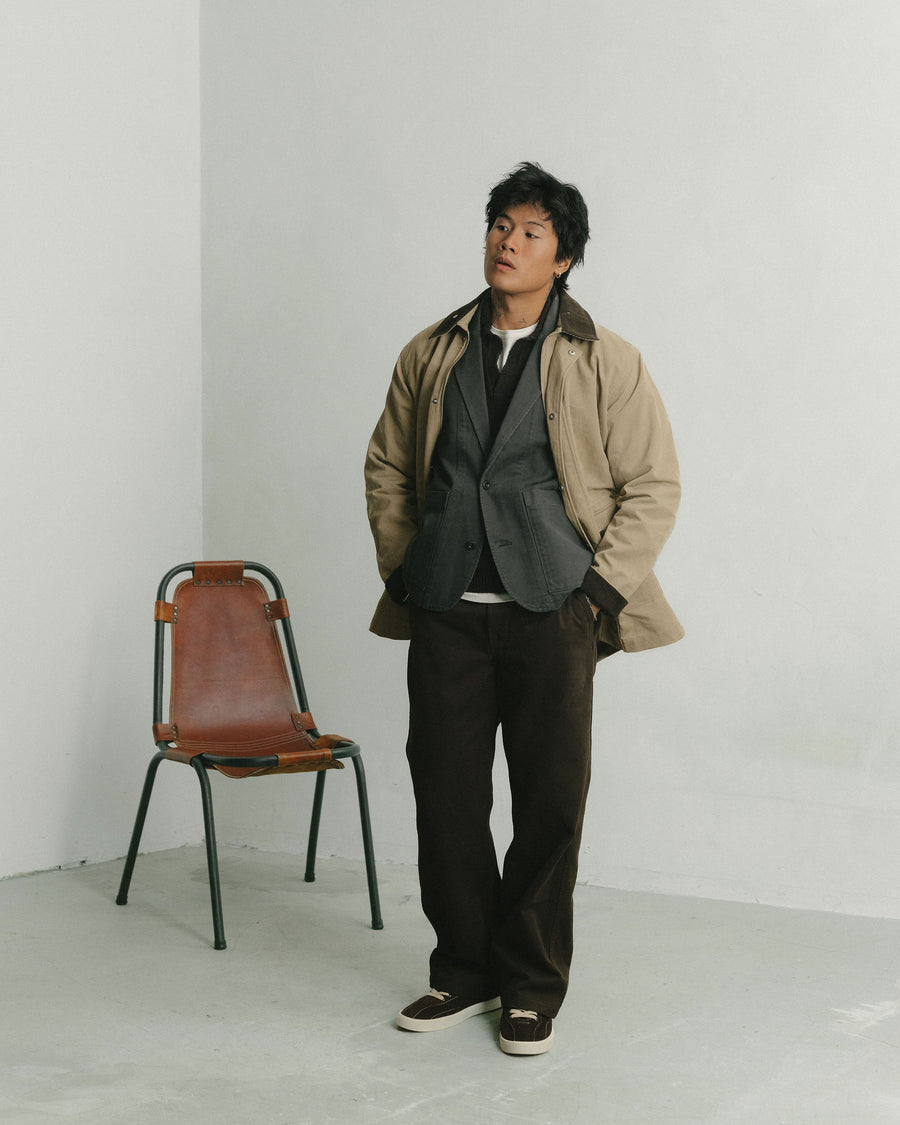 A man in a tan waxed jacket and brown pants stands in front of a leather chair.