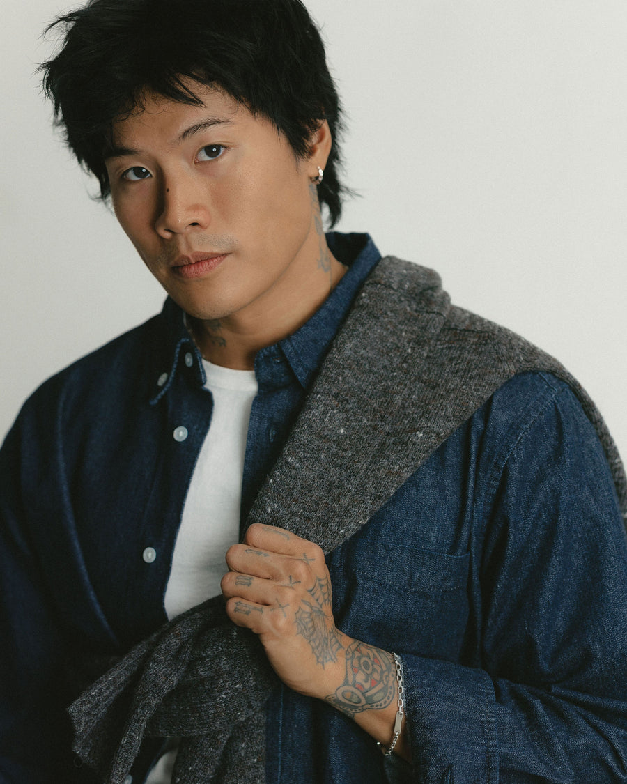 A close up portrait of a man wearing a dark denim shirt and a sweater over his shoulder.