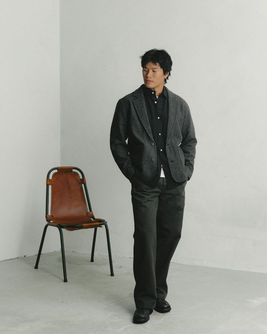 A man in a wool blazer layered over a black button down shirt poses in front of a leather chair.