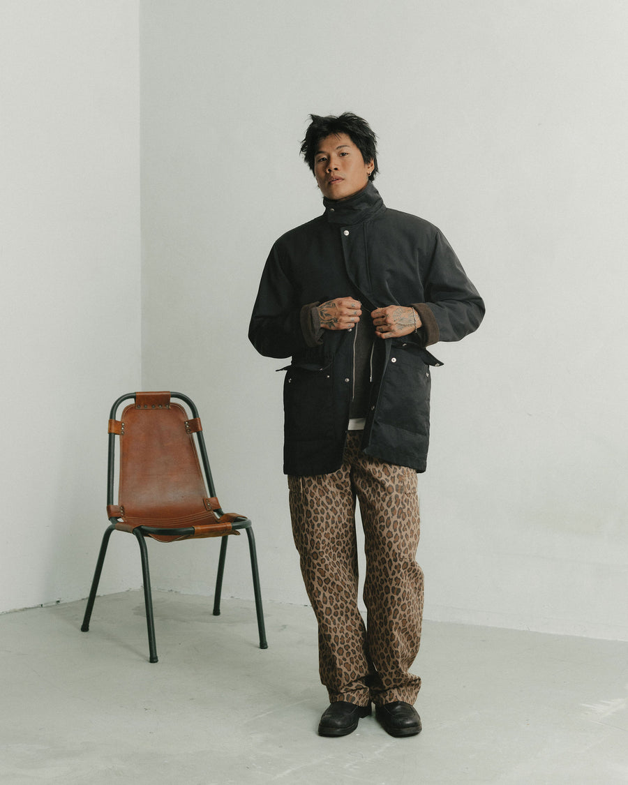 A man in a black nylon coat over leopard print pants stands and buttons the coat up.