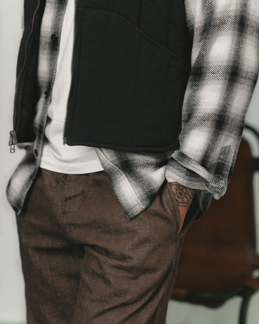 A detail shot of the textures of a black and white flannel worn open under a black nylon vest, paired with brown denim pants.