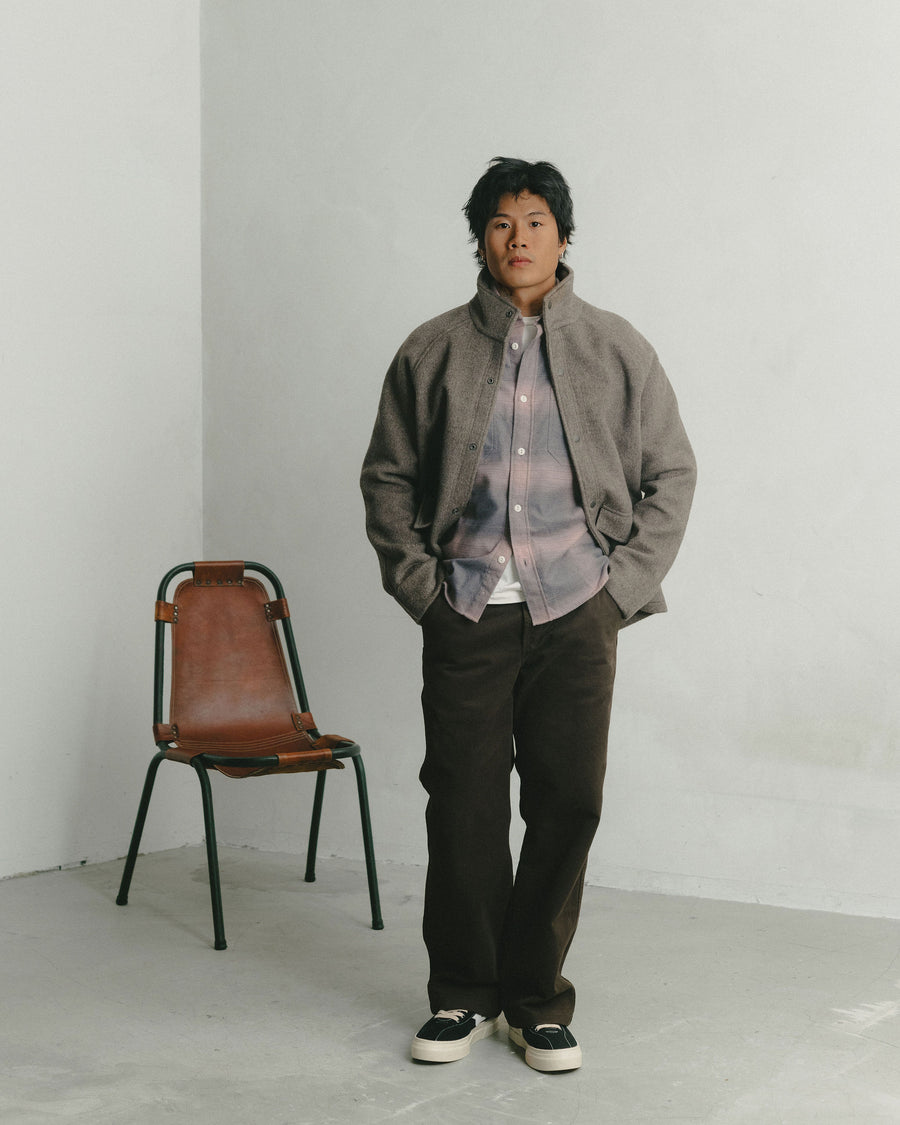 A man in a pink flannel layered under a tan wool coat poses with his hands in his pockets.