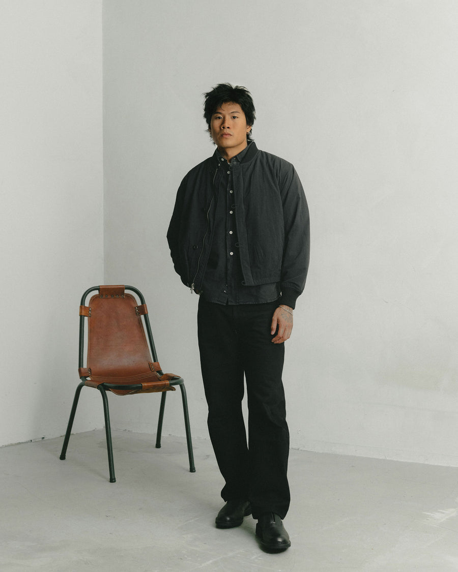 A man in a black cropped bomber jacket and black jeans stands in front of a leather chair.