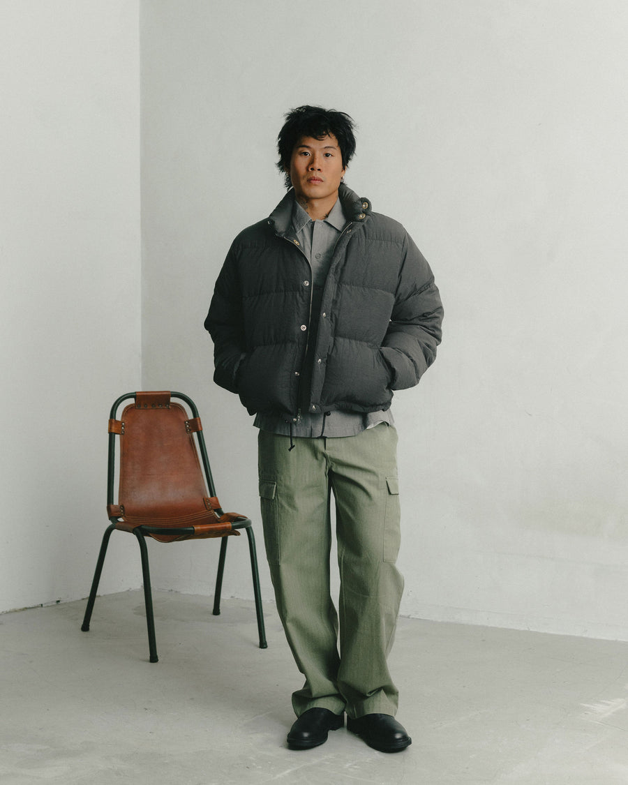 A man in a black nylon down jacket and olive cargo pants stands in front of a leather chair.