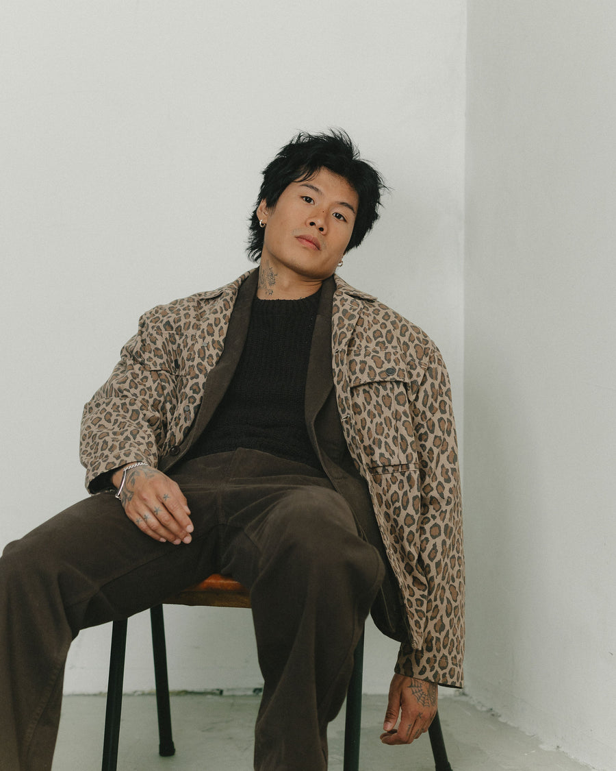 A man in a leopard print jacket layered over a chocolate brown cotton suit sits in a relaxed, effortless devil-may-care style in a leather chair.