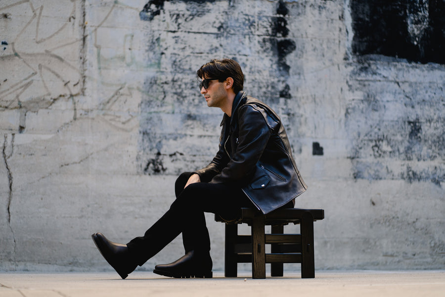 A man in a leather perfecto jacket sits on a low wooden stool.