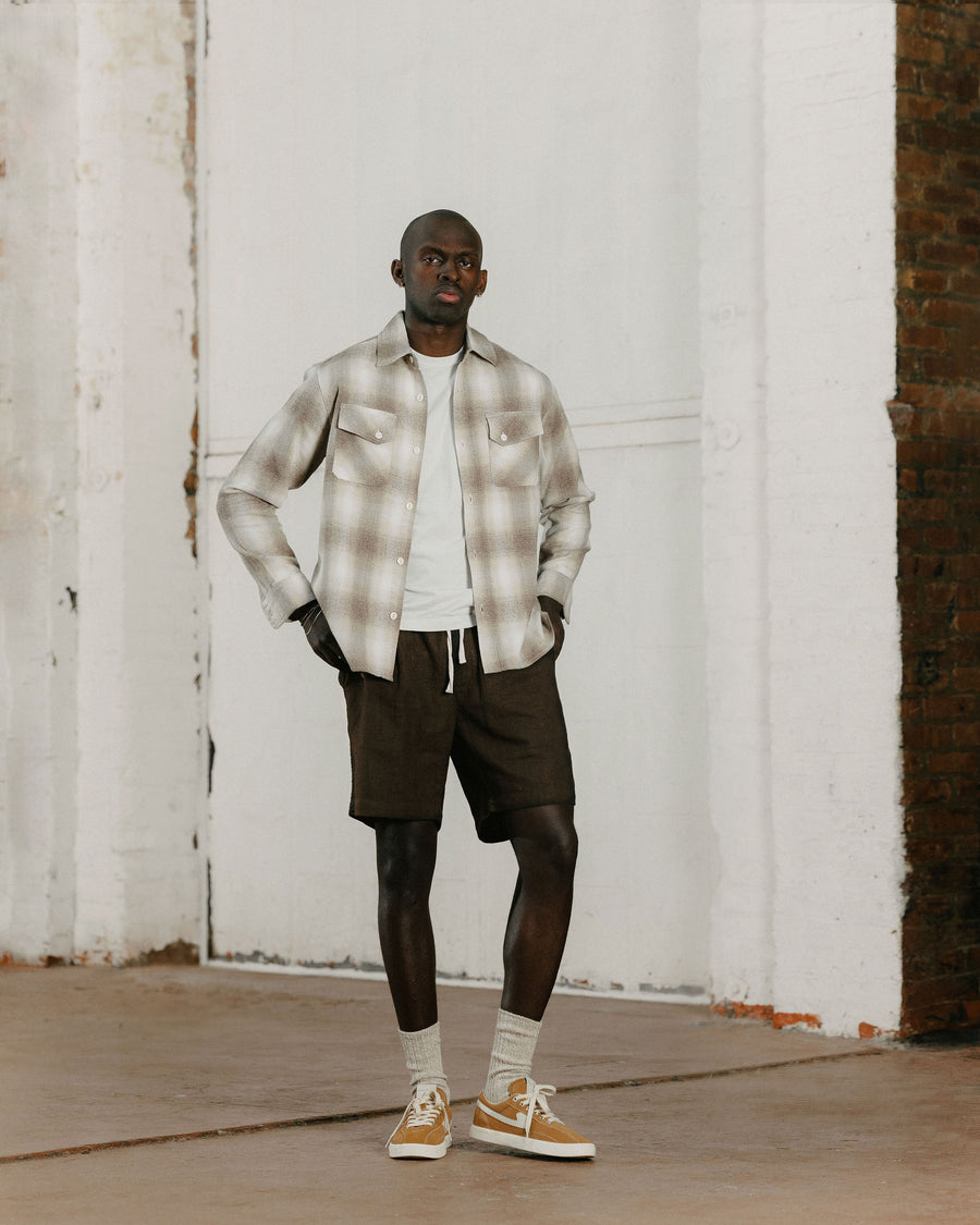 A man in a tan plaid flannel and brown shorts poses.