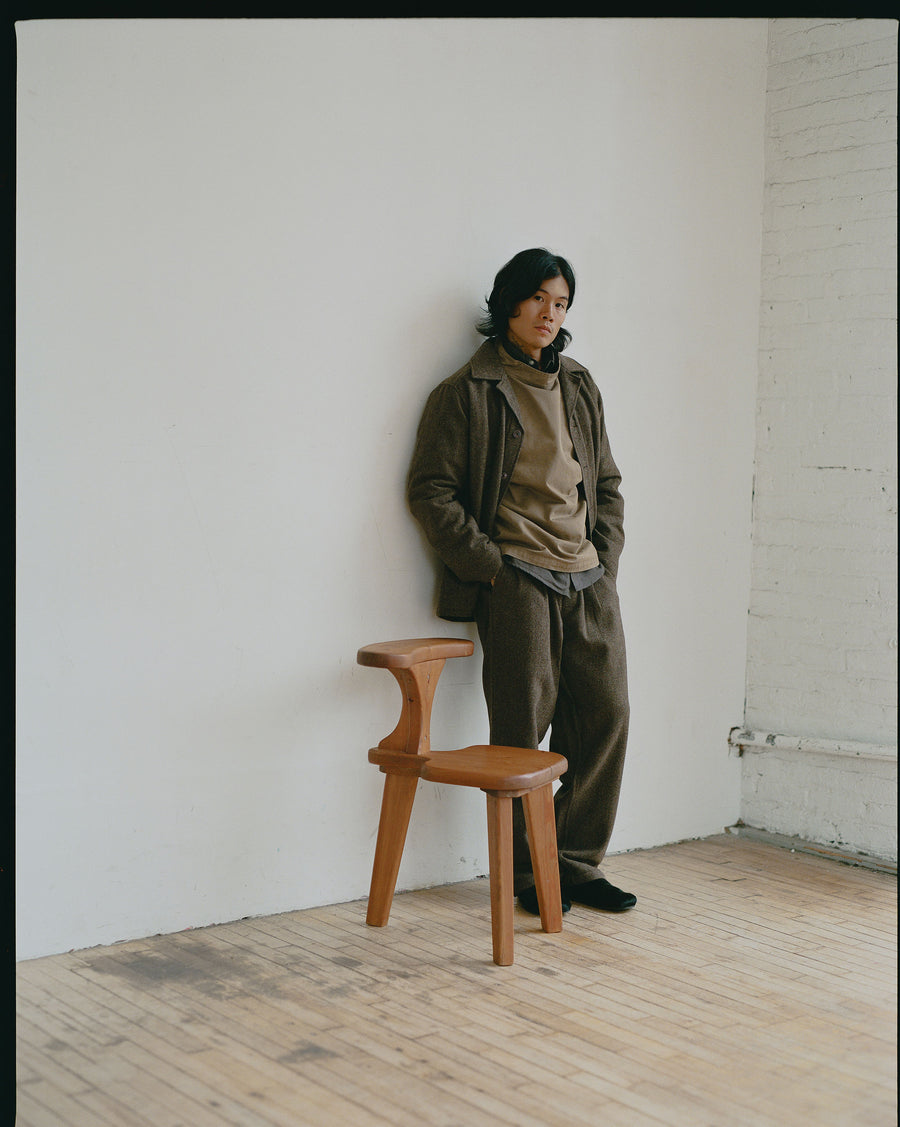 A man in a tan pullover layered under an earth toned wool coat leans against a wall.