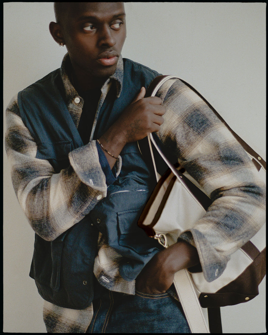 A man in a vest layered over a flannel carries a tan and brown bag.