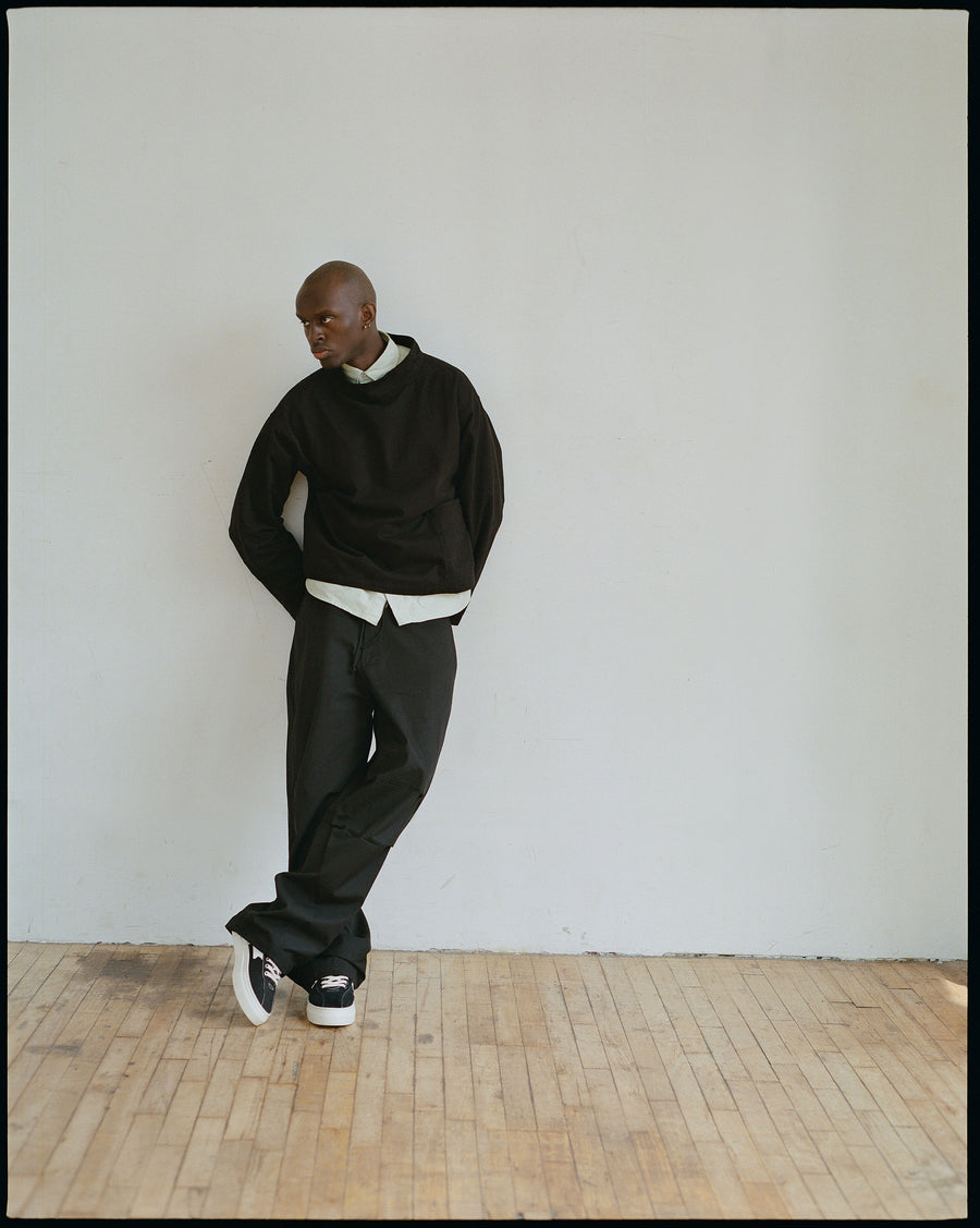 A man in a black smock layered over a button down shirt and big flowy black pants leans against a wall.