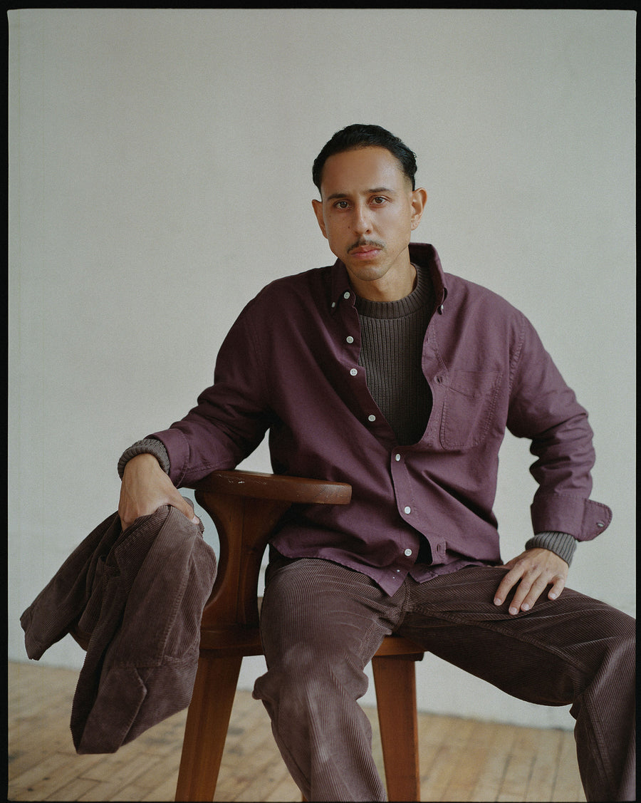 A man in a burgundy shirt and corduroy suit sits in a chair.