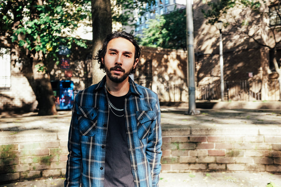 A man stands in a brightly lit park wearing a blue 3sixteen flannel open over a black tee.