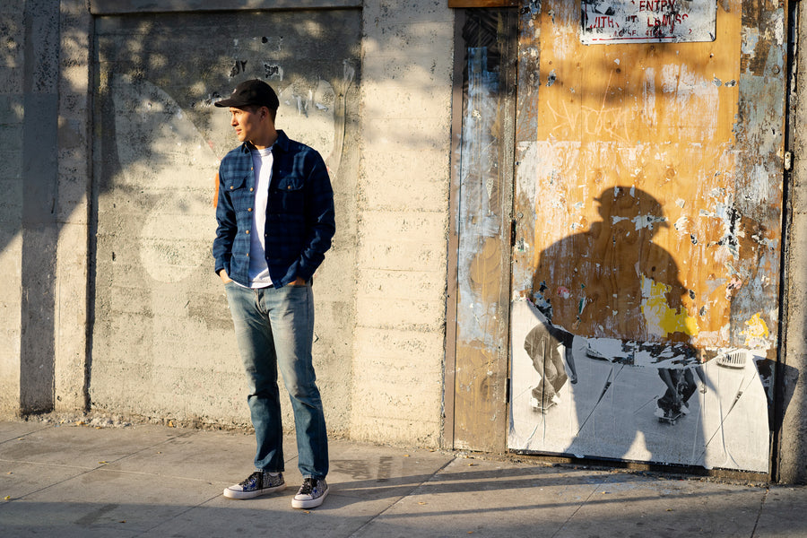 A man stands on a street in a blue flannel.