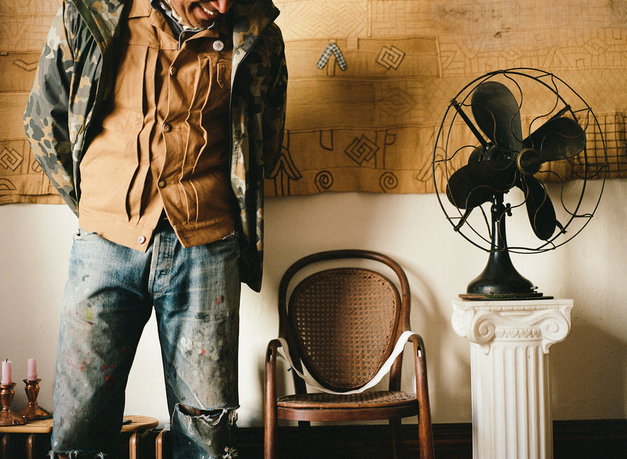 A man in a camoflauge jacket and worn in jeans stands by a chair and a fan.