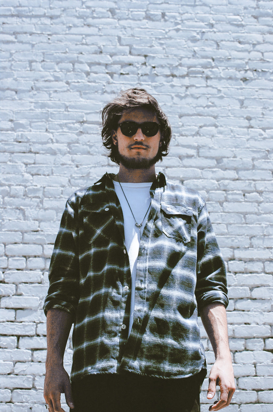 A man in a blue plaid check flannel and sunglasses in front of a white brick wall.