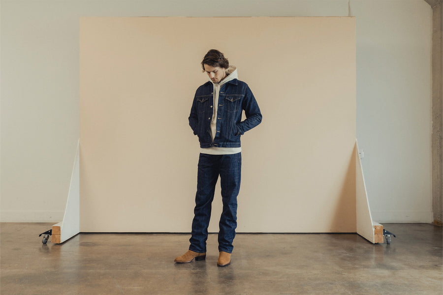 A man in a denim jacket, hoodie and jeans stands in front of a rolling backdrop.