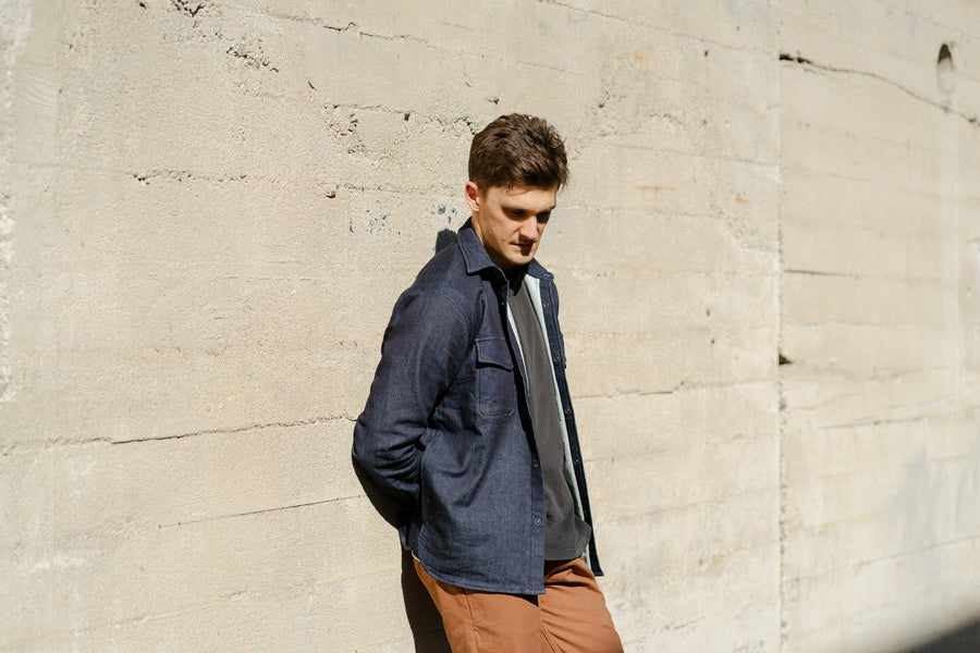 A man in a blue shirt and rust colored chinos leans up against a wall.