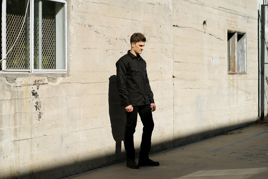 A man in a black shirt stands against a wall.