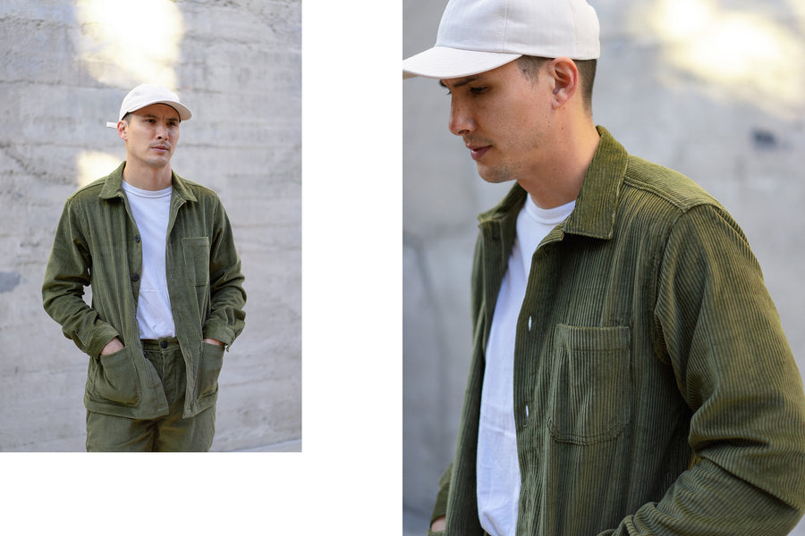 A young man wears a natural baseball cap and a corduroy shop jacket in olive.