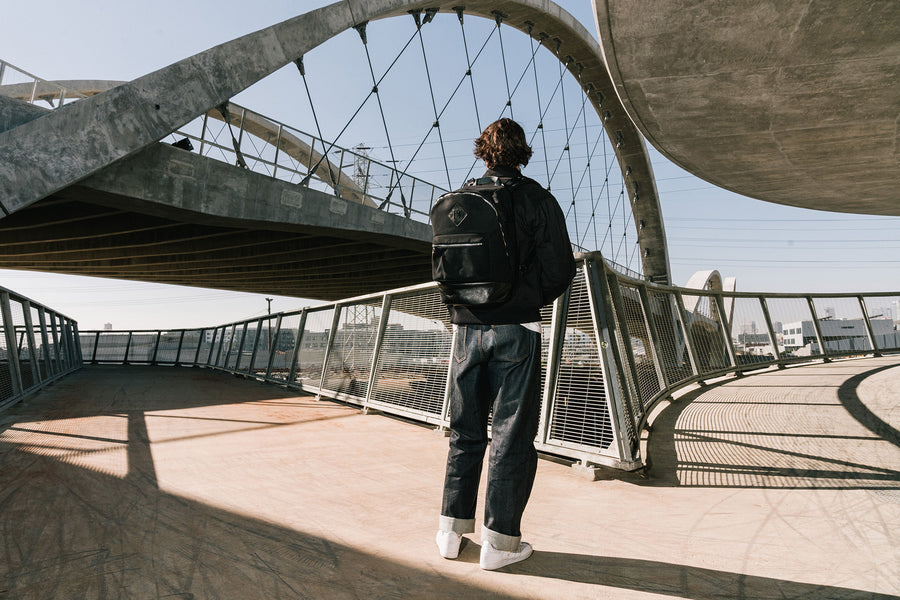 A man in a black jacket and a black backpack stands on a bridge.